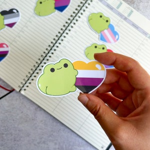 May include: A set of frog stickers with pride flags. The stickers feature a green frog holding a heart-shaped sticker with the colours of the genderqueer pride flag. The other stickers show a frog with a transgender pride flag and a frog with a bisexual pride flag.