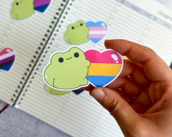 May include: A set of frog stickers with different pride flags. The stickers are green with a white outline and feature a frog holding a heart-shaped flag. The flags include the bisexual, transgender, and pansexual flags.