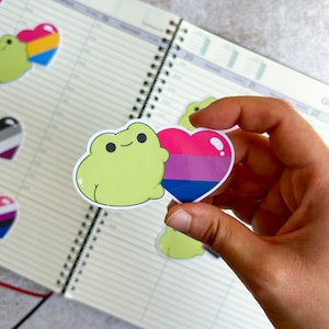 Pode incluir: Um adesivo de sapo verde segurando um adesivo em forma de coração com as cores da bandeira do orgulho bissexual. O coração é rosa, roxo e azul.