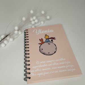 May include: A pink spiral notebook with a quote from The Little Prince in white text. The quote reads: "Ci sara sempre un altra opportunita, un altra amicizia, un altro amore, una nuova forza. Per ogni fine, c'e un nuovo inizio." The notebook has a cartoon illustration of The Little Prince on a planet with a fox.