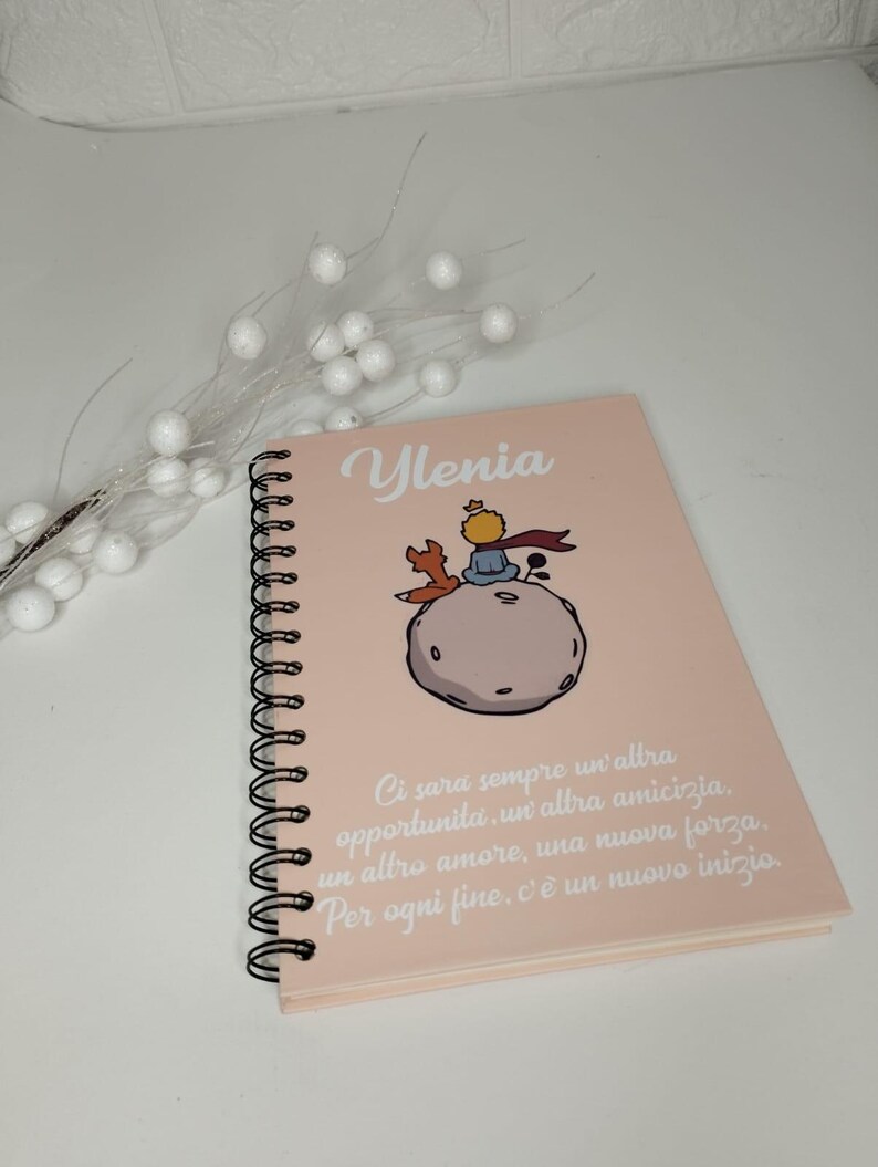 May include: A pink spiral notebook with a quote from The Little Prince in white text. The quote reads: "Ci sara sempre un altra opportunita, un altra amicizia, un altro amore, una nuova forza. Per ogni fine, c'e un nuovo inizio." The notebook has a cartoon illustration of The Little Prince on a planet with a fox.