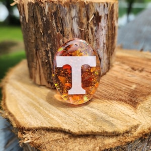 May include: A white letter "T" is embedded in a clear resin charm with orange, gold, and iridescent glitter. The charm is sitting on a piece of wood.