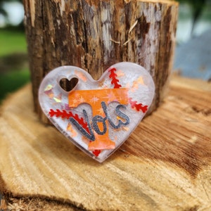 Może przedstawiać: Biały, sercowaty brelok z żywicy z błyszczącym pomarańczowym i białym "T" oraz napisem "Vols" w srebrnym brokacie. Tło zdobią czerwone i białe szwy baseballowe.