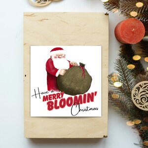 Op de afbeelding: Een vierkante wenskaart met een cartoonillustratie van de Kerstman in een rood pak die een grote, getextureerde donkergroene zak vasthoudt. De tekst "Have a MERRY BLOOMIN' Christmas" is gedrukt in een mix van zwarte en rode letters.