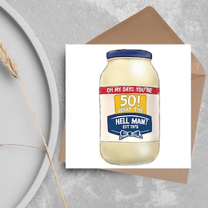 Może przedstawiać: Kartka z życzeniami z zabawną ilustracją słoika majonezu. Tekst na kartce brzmi: "OH MY DAYS YOU'RE 50! WHAT THE HELL MAN? EST 1975". Kartka leży na brązowej kopercie.