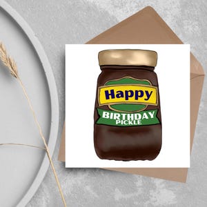 May include: A birthday card with a cartoon illustration of a pickle jar. The jar has a gold lid and a label that says "Happy Birthday Pickle" in yellow and green. The card is on a brown envelope.