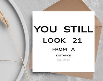 Peut inclure: Une carte de vœux blanche avec le texte "YOU STILL LOOK 21 FROM A DISTANCE HAPPY BIRTHDAY" en noir, posée sur une enveloppe marron. La carte est sur une surface grise avec un élément décoratif.