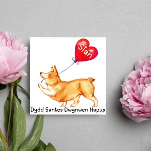 May include: A square print featuring a watercolor illustration of a corgi dog holding a red heart-shaped balloon with the name "Sian" on it. The dog is light brown and the balloon is connected with a blue string. The text "Dydd Santes Dwynwen Hapus" is at the bottom.