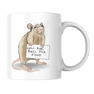 May include: A white ceramic mug featuring a cartoon illustration of a tan rat standing on its hind legs. The rat holds a white rectangular sign with the black handwritten text "WILL RING BELL FOR FOOD" printed on the front of the mug.