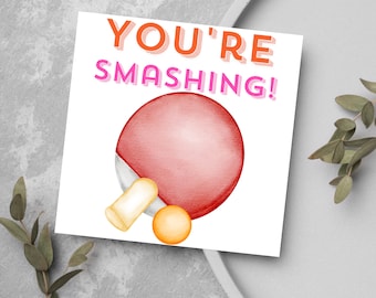 Op de afbeelding: Een witte kaart met de tekst "YOU'RE SMASHING!" in oranje en roze letters. Een roze en witte pingpongbat en een gele pingpongbal zijn geïllustreerd op de kaart.