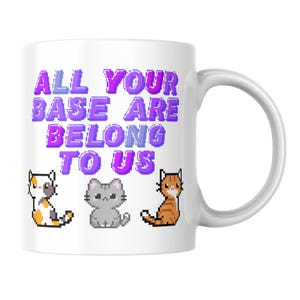 May include: A white ceramic mug featuring the purple pixelated text ALL YOUR BASE ARE BELONG TO US. Below the text are three pixel art cats: a calico cat on the left, a grey cat in the centre, and an orange tabby cat on the right side.