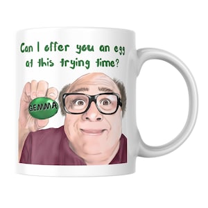 May include: A white ceramic mug featuring a printed illustration of a person with glasses wearing a maroon shirt. The person holds a green egg labelled GEMMA. Above the figure, green text reads: Can I offer you an egg at this trying time?