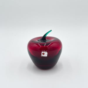 May include: A handmade Blenko glass apple sculpture. The apple is a deep red colour with a dark base and a green stem. The Blenko logo and "HANDMADE" are visible on the front of the apple.