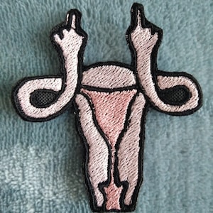 Può includere: Toppa ricamata rosa e nera di un utero con due braccia alzate in aria che fanno il dito medio.