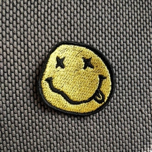 Può includere: Toppa ricamata con smiley giallo, contorno nero, occhi a croce e lingua. La toppa è posizionata su uno sfondo grigio strutturato. Perfetta per personalizzare abbigliamento e accessori.