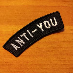Op de afbeelding: Zwarte stoffen patch met witte geborduurde tekst die "ANTI-YOU" luidt.
