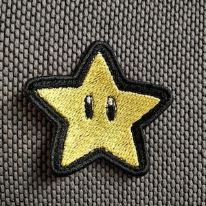 Può includere: Una toppa a forma di stella gialla con contorni neri e due occhi neri. La stella è ricamata con filo giallo e gli occhi sono ricamati con filo nero.