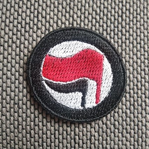 Puede incluir: Parche bordado en blanco y negro con una bandera roja en el centro. La bandera está estilizada para parecer un puño que sostiene un martillo y una hoz.