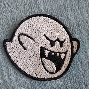 Könnte beinhalten: Ein gestickter Aufnäher mit einem Cartoon-Geist mit schwarzem Umriss und einem breiten, zahnigen Grinsen. Der Geist hat ein einzelnes Auge und zwinkert.