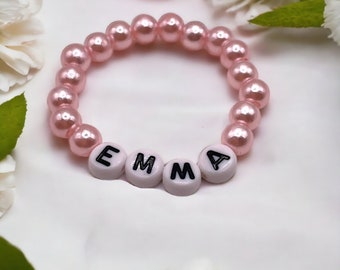 May include: A pink beaded bracelet with the name "EMMA" spelled out in white letter beads. The bracelet is surrounded by white and pink flowers and green leaves, creating a soft, feminine aesthetic.