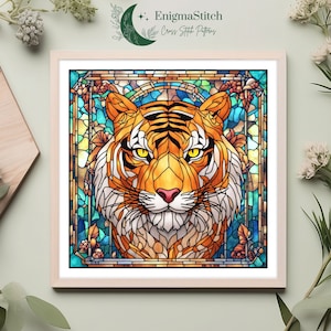 May include: Framed cross-stitch pattern featuring a tiger's face in a stained-glass style. The tiger has orange and black stripes, yellow eyes, and a pink nose. The background is a mix of blue, green, and yellow. The frame is light wood, and the text "EnigmaStitch Cross Stitch Patterns" is at the top.
