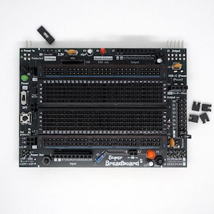 Puede incluir: Una placa de circuito impreso negra Super Breadboard 2 que presenta un área central con filas numeradas y columnas con letras, varios componentes electrónicos, un puerto de alimentación USB-C y cabezales de pines.