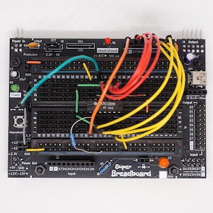 Puede incluir: Una Super Breadboard negra con varios componentes electrónicos y cables de colores. La placa presenta secciones etiquetadas para alimentación, entrada y salida, junto con un indicador de 'Cortocircuito'. El texto en la placa incluye 'N74LS393N' y 'Super Breadboard'.