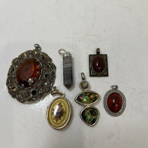 May include: A collection of six assorted pendants including a large oval filigree piece with an amber-colored stone, a gray striped crystal point, a rectangular pendant with a dark red stone, a floral oval locket, and two iridescent shell pieces.