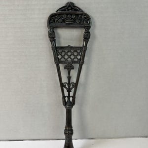 May include: A dark metallic, elongated handheld tool featuring an intricate openwork design. The top section displays a floral motif within a rectangular frame, followed by a lattice pattern and a slender, tapered handle with a rounded base.