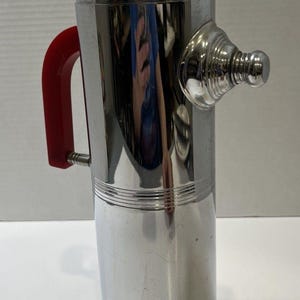 May include: A vintage chrome cocktail shaker with a red handle and top. The shaker has a cylindrical body with a spout and a base. The handle and top are a vibrant red, contrasting with the shiny chrome finish. A classic barware item.