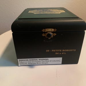 May include: A dark green wooden cigar box with a gold-colored metal latch. The front features the text 20 - PETITE ROBUSTO 54 x 4 1/4 and a Surgeon General warning label. A gold-colored decorative label is visible on the top lid.