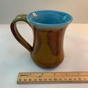 May include: A handmade ceramic mug with a brown exterior, a blue interior, and a curved handle. The mug has a capacity of approximately 12 ounces. The mug is 4 inches tall and 3 inches wide.