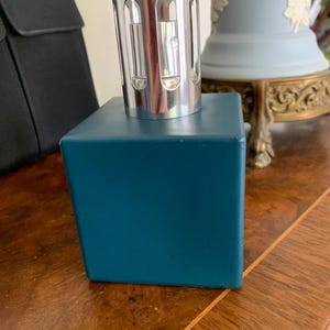 May include: A teal-colored, cube-shaped fragrance diffuser with a silver metal top. The diffuser sits on a wooden surface. The top has a series of vertical openings. A black object is visible in the background.