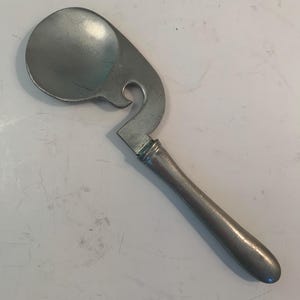 May include: A vintage, silver-toned ice cream scoop with a distinctive design. The scoop features a rounded bowl, a curved, open-sided section, and a long, cylindrical handle. The metal has a slightly aged appearance.