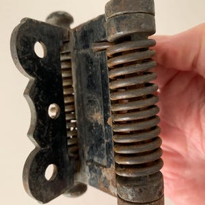 May include: An antique black metal hinge with a spring mechanism. The hinge has a decorative, scalloped edge and a cylindrical spring. The metal shows signs of age and wear, with a rusty appearance.