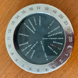 May include: A silver-colored world time clock with a black face. The clock face has a rotating disc with city names and time zones. The outer ring of the clock has numbers 1 through 24.