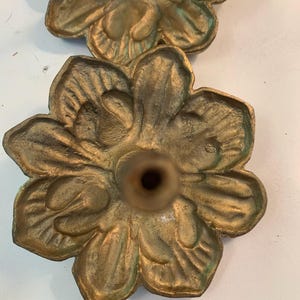 May include: Two brass-colored metal drawer knobs shaped like six-petaled flowers. Each knob features a textured, organic petal design with a central cylindrical post extending outward. The metal has a weathered, aged finish with subtle green oxidation.