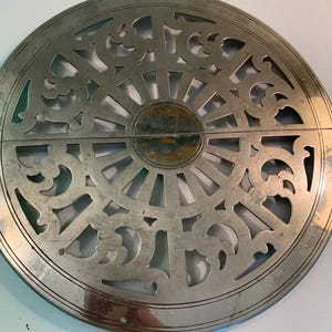 May include: Circular silver-toned metal trivet with an intricate openwork design. The trivet features a central sunburst pattern surrounded by decorative scrollwork. The metal has a slightly aged appearance, suggesting it is vintage or antique.