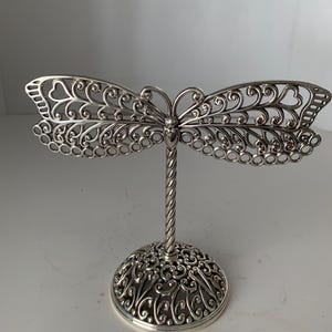 May include: A silver-colored dragonfly-shaped jewelry display stand. The wings feature an openwork design with heart and swirl patterns. The stand has a twisted stem and a decorative base with intricate scrollwork.