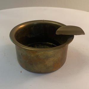 May include: A round, metallic ashtray with a tarnished brass finish. The vessel features a deep, cylindrical basin and a single, wide, curved rest extending from the rim, designed to hold a cigarette or cigar securely in place.