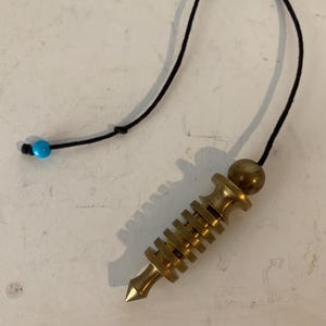 May include: A brass pendulum with a pointed tip and a series of horizontal ridges. It is suspended by a black cord with a small blue bead at the end. The pendulum has a gold-colored metal bead at the top.