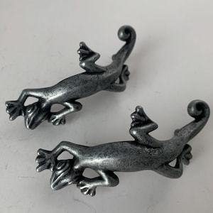 May include: Two pewter-colored lizard-shaped cabinet pulls. Each lizard has detailed features, including textured skin and curled tails. The pulls are designed to be mounted horizontally and add a unique decorative element to furniture.