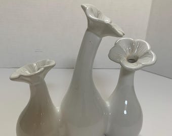 May include: A white ceramic vase with three flower-shaped openings. The vase has a glossy finish and a unique design, perfect for displaying flowers or as a decorative piece. The vase is a bright white color.