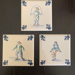 May include: Three vintage ceramic tiles with blue and white floral designs and hand-painted scenes of people in traditional clothing. The first tile shows a person jumping rope, the second tile shows a person wearing a hat and green jacket, and the third tile shows a person riding piggyback on another person.