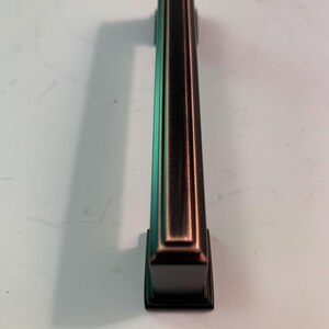 May include: A bronze-colored cabinet pull with a rectangular design. The pull has a dark finish with copper-colored accents and a square base. The handle is vertically oriented, with a long, rectangular grip.