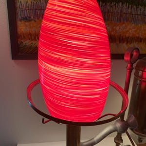 May include: A cylindrical glass lamp shade featuring a vibrant red and white swirled pattern. The shade is illuminated from within, creating a bright glow. It is mounted on a metallic base with a circular support ring and a vertical central post.