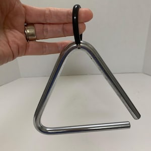 May include: A shiny, silver-colored triangle musical instrument with a black handle. The triangle is held by a hand, showcasing its polished metal finish and simple design. This percussion instrument is used in orchestras and bands.