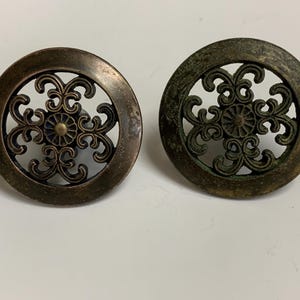 May include: Two antique bronze cabinet knobs with intricate openwork designs. Each knob features a circular frame with a detailed floral pattern in the center. The knobs have a vintage, aged appearance, suggesting they are repurposed or vintage.