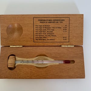 May include: A wooden box with a thermometer inside. The thermometer has a wooden handle shaped like a barrel. The box has a label that reads "Temperatures Conseillees Pour Le Service Du Vin" and lists different types of wine and their recommended serving temperatures in degrees Celsius.
