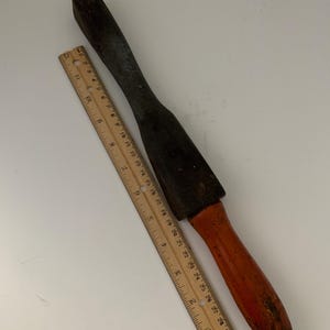 May include: A wooden and metal tool with a long, rectangular handle and a flat, rectangular head. The handle is painted orange and the head is black. The tool is about 12 inches long.